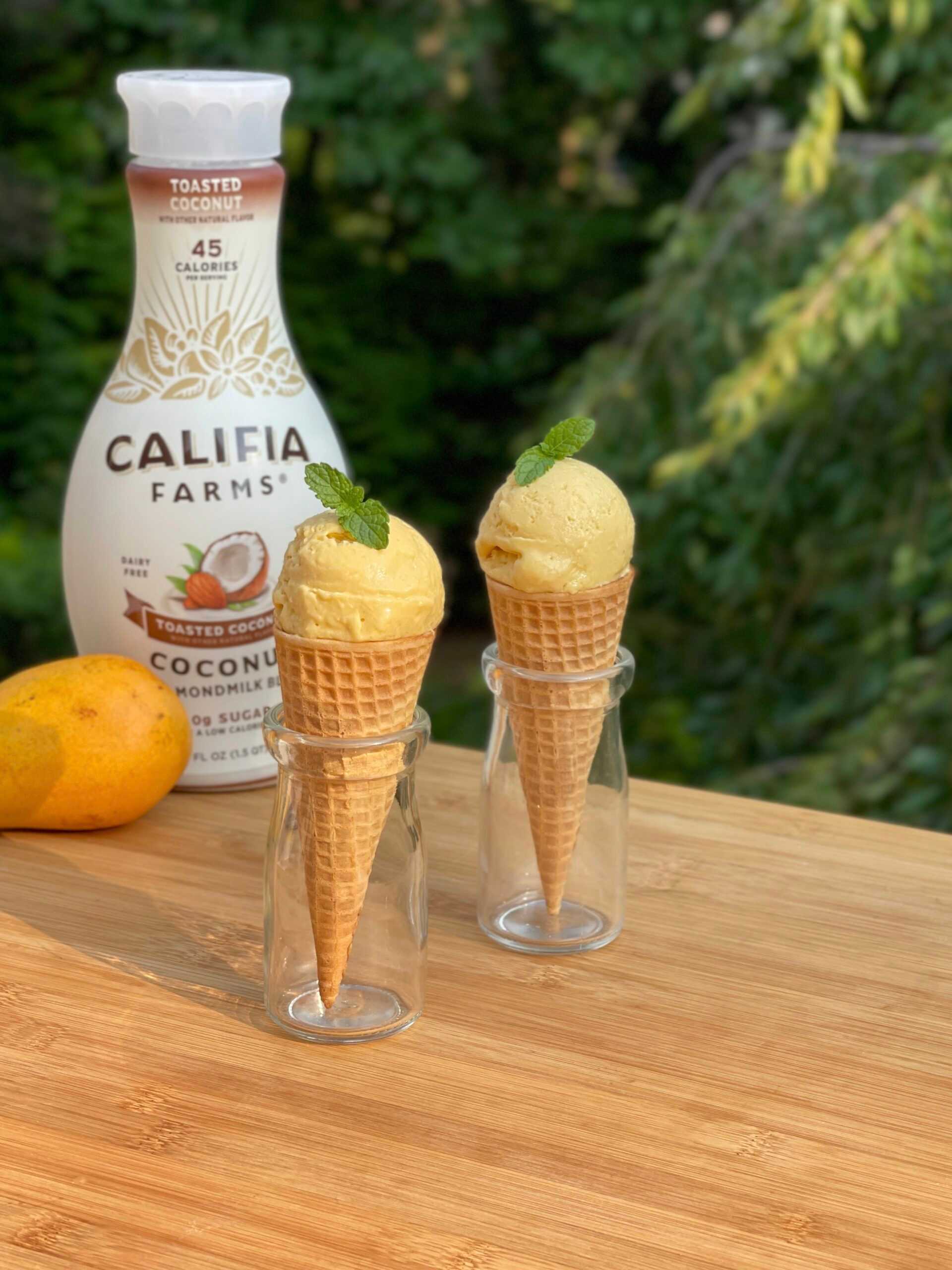 Small cones with scoops of mango ice cream sit at the center of the image, with a bottle of Califia Farms Toasted Coconut Almondmilk in the background.