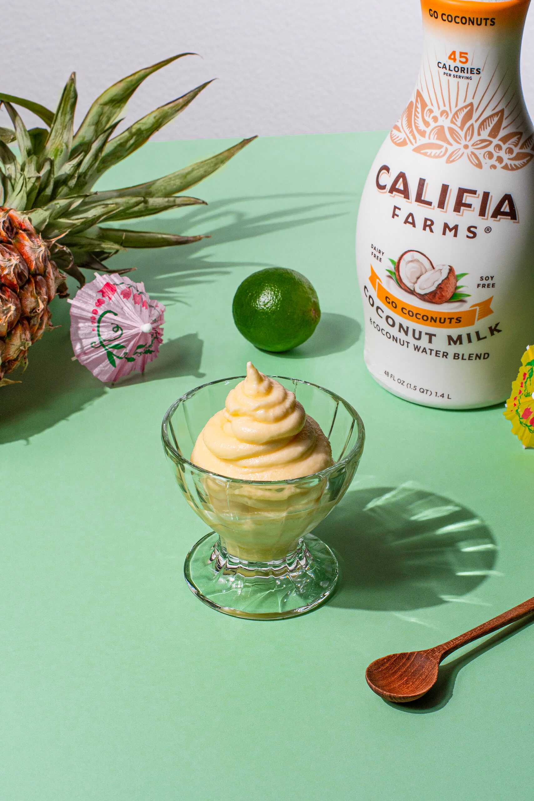 A small portion of Pineapple Coconut Soft Serve sits in the center of the image, surrounded by a pineapple, lime, wooden spoon, and Califia Farms Go Coconuts Coconutmilk.