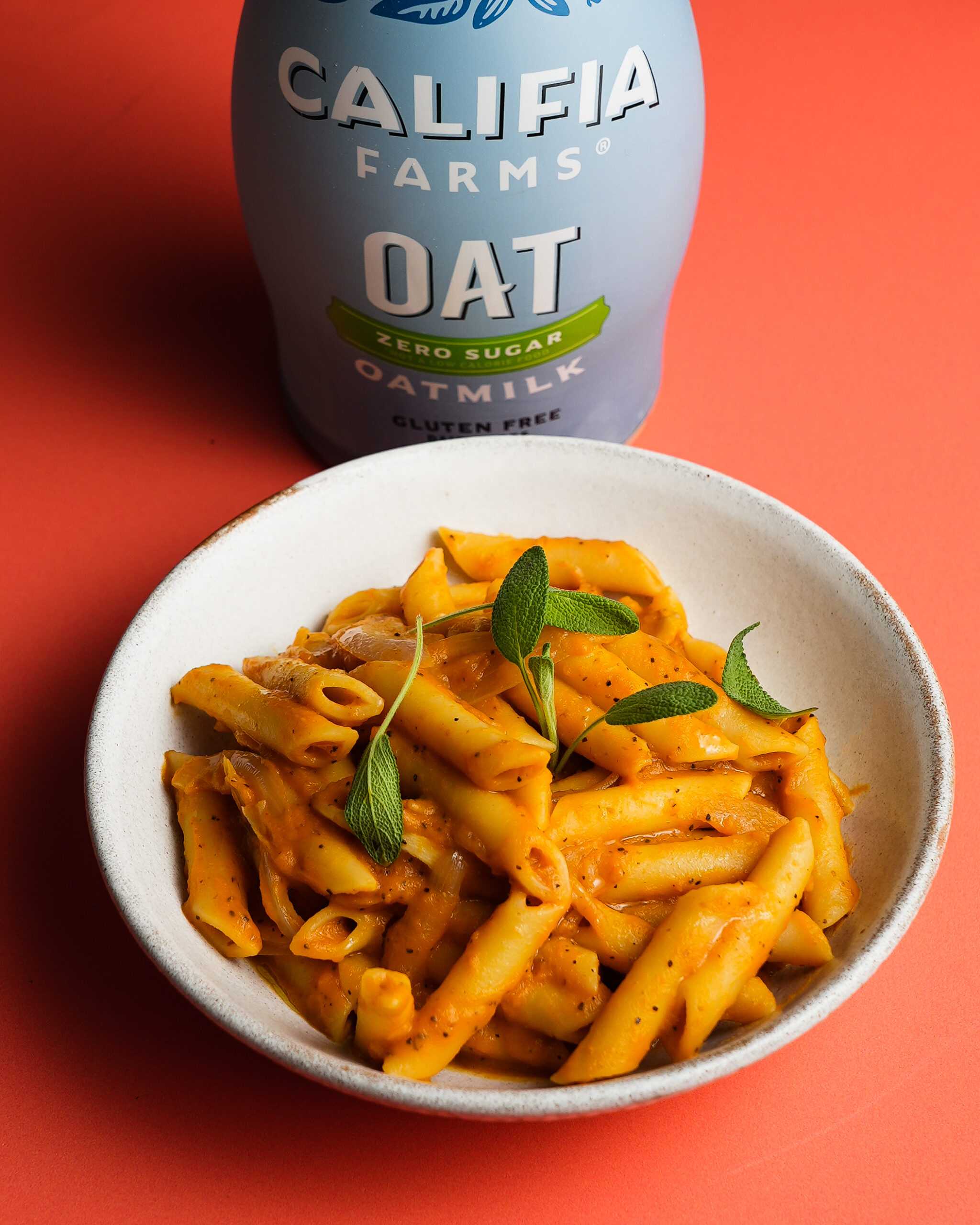 A bowl of pumpkin pasta sits in the foreground of the image, with Califia Farms Zero Sugar Oatmilk behind.