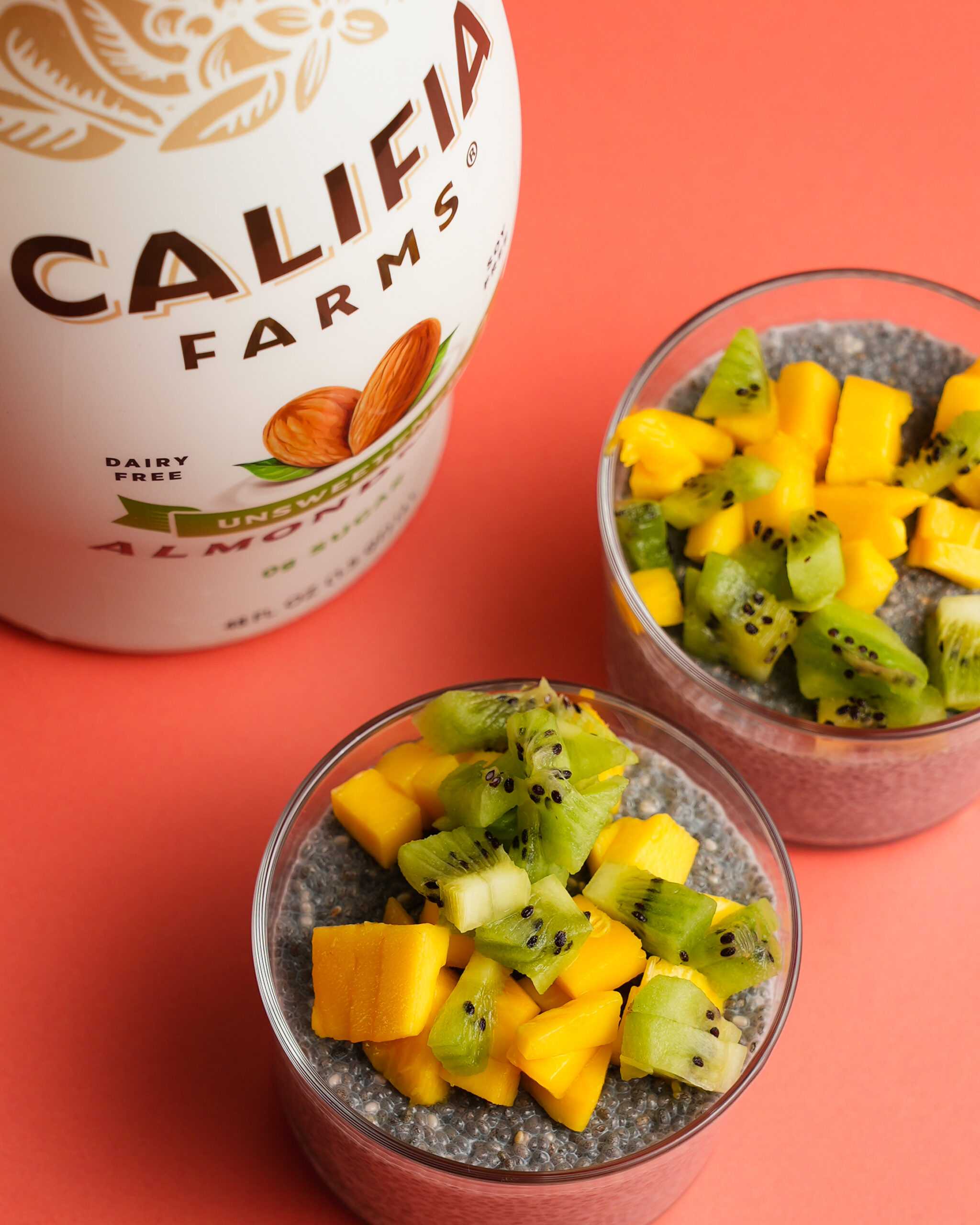Two chia seed puddings with mango and kiwi on top sit in the front of the image, with Califia Farms Unsweetened Almondmilk off to the side.