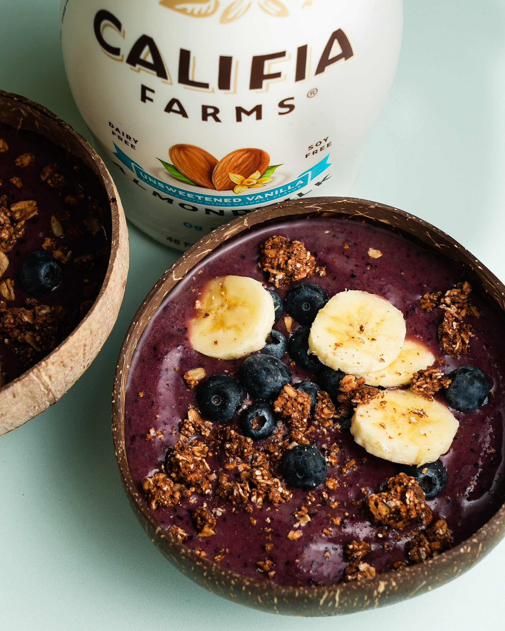 Acai bowl with chopped banana, blueberries, and granola sits in the front of the image, with Califia Farms Unsweetened Vanilla Almondmilk in the back.