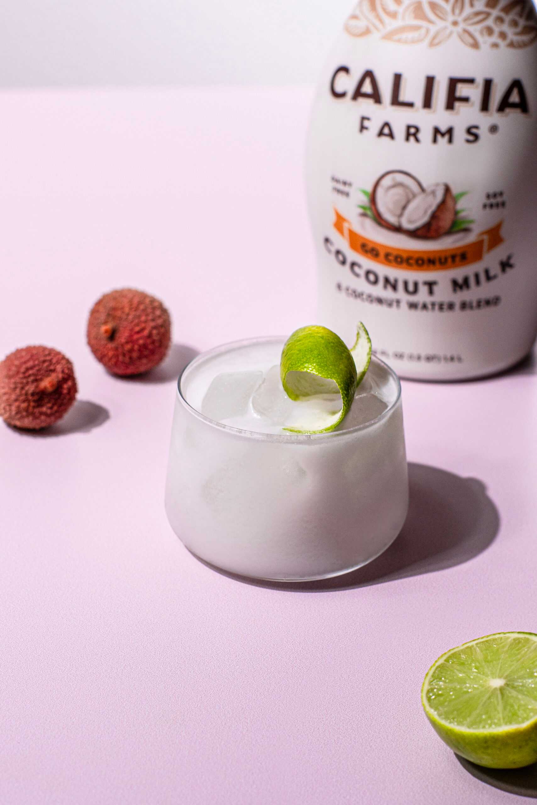 Coconut lychee cocktail garnished with a lime wedge sits in the center of the image, with lychee fruit off to the side and Califia Farms Go Coconuts Coconutmilk in the background.