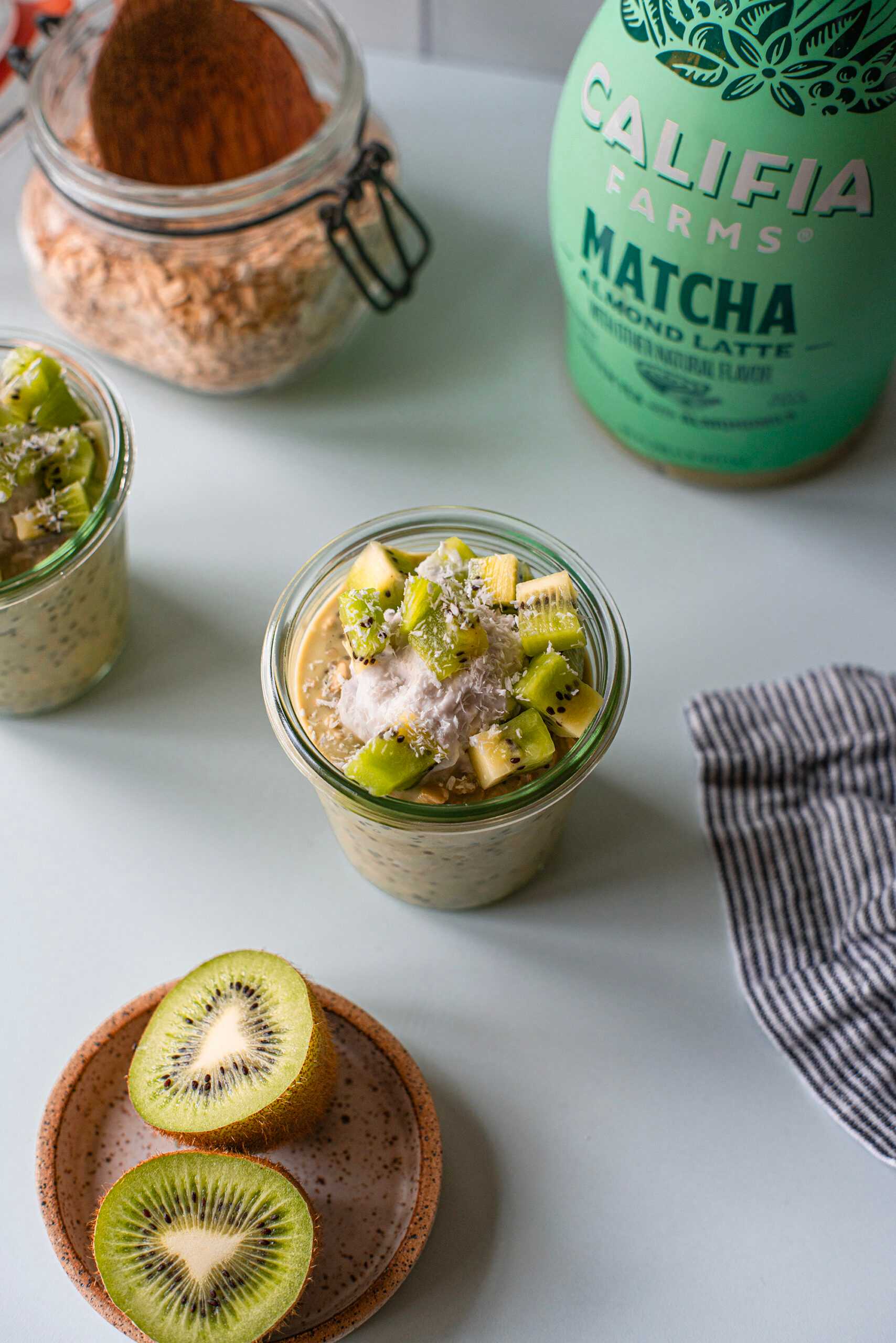 A jar of Matcha Overnight Oats sits at the center of the image, with a bowl of kiwi fruit nearby and Califia Farms Matcha Latte in the back of the image