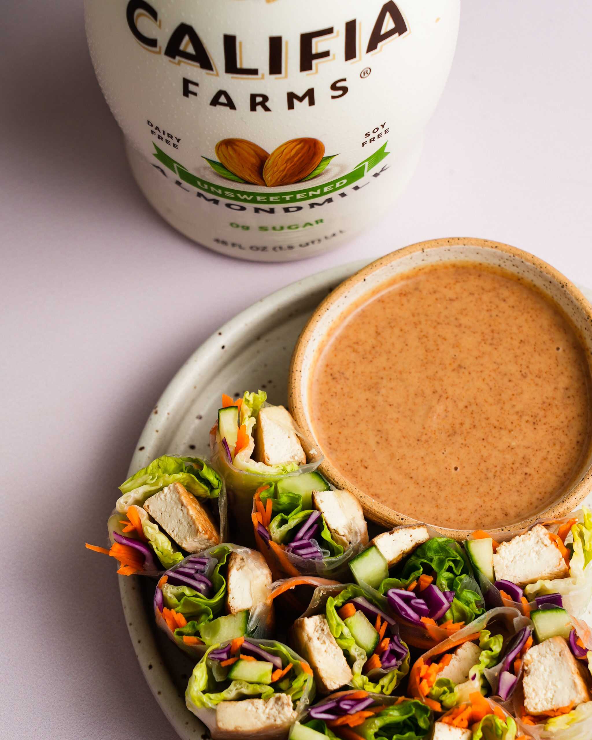 Bright colored vegetable spring rolls sit in the center of the image with almond butter dipping sauce in a separate container. Califia Farms Unsweetened Almondmilk sits in the back of the image.