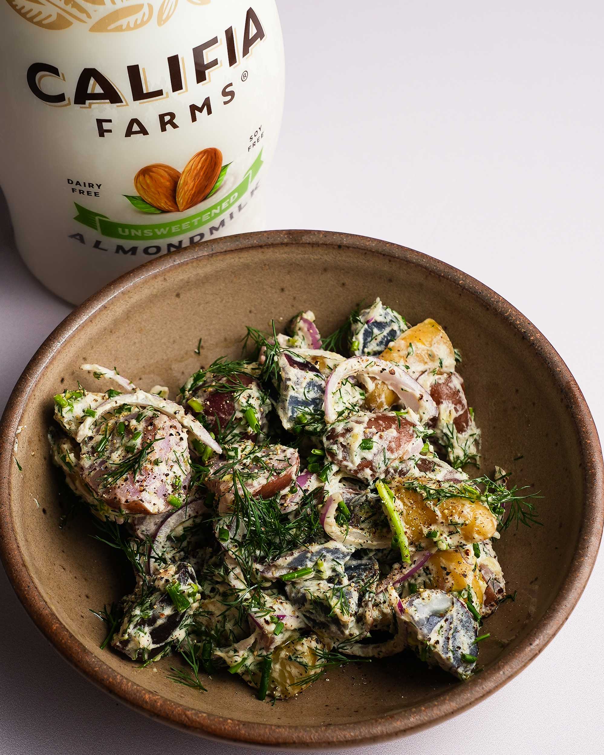 A bowl of potato salad sits in the center of the image, with Califia Farms Unsweetened Almondmilk behind.