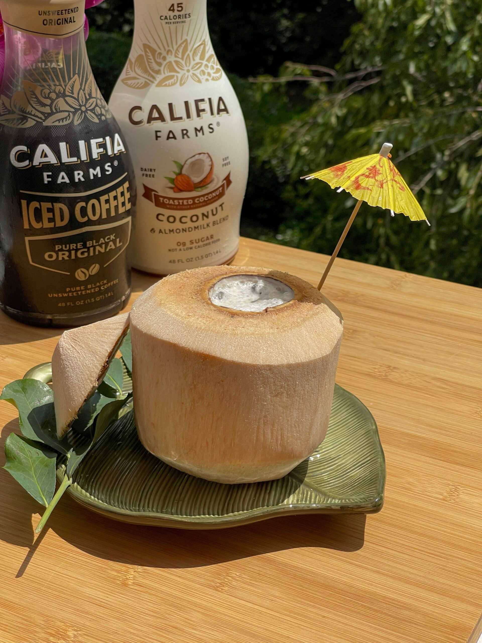 A frothy iced coffee sits inside a coconut at the front of the image, with Califia Farms Iced Coffee and Toasted Coconut Almondmilk behind.